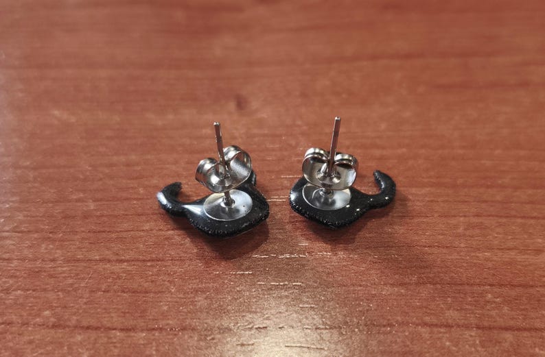 May include: A pair of black stud earrings with a mustache design. The earrings have a silver post and backing. The earrings are on a brown wooden surface.