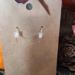 May include: A pair of gold earrings with clear plastic backs are displayed on a brown earring card. The earrings are hanging from the card. The background includes a pumpkin and autumn leaves.