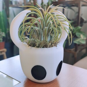 May include: A white ceramic planter shaped like the Knight from the video game Hollow Knight, with a green air plant growing inside.