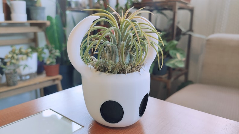 May include: A white ceramic planter shaped like the Knight from the video game Hollow Knight, with a green air plant growing inside.