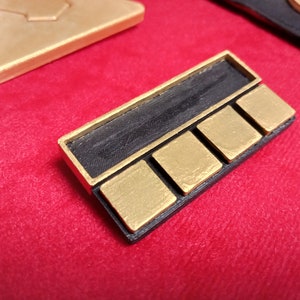May include: A black and gold rectangular badge with four gold squares and a black rectangular space above the squares.