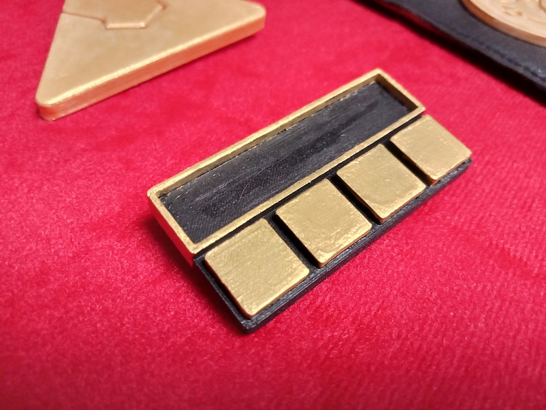 May include: A black and gold rectangular badge with four gold squares and a black rectangular space above the squares.