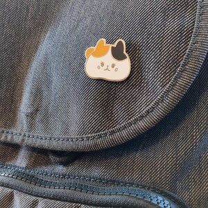 May include: A white enamel pin with a cartoon cat design. The cat has orange, black, and white fur and a cute expression. The pin is on a dark gray fabric background.