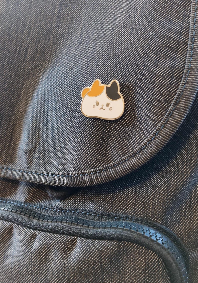 May include: A white enamel pin with a cartoon cat design. The cat has orange, black, and white fur and a cute expression. The pin is on a dark gray fabric background.