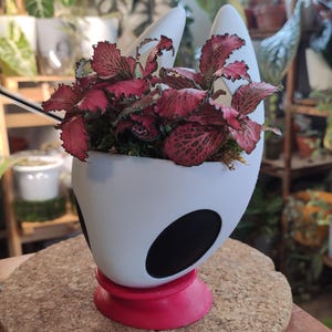 May include: White ceramic planter shaped like a knight's helmet with black accents. A red base supports the planter. The planter is filled with a pink and green leafy plant.