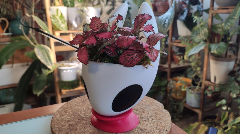 May include: White ceramic planter shaped like a knight's helmet with black accents. A red base supports the planter. The planter is filled with a pink and green leafy plant.
