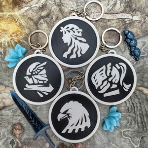May include: A collection of black and white keychains with various graphic designs, including a profile, an eagle, and other stylized figures. Each keychain has a silver ring. Decorative blue objects and a blue sword are also present.