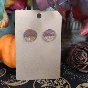 May include: A pair of round wooden earrings with gold flakes embedded in clear resin. The earrings are displayed on a brown card.