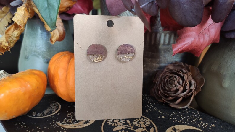 May include: A pair of round wooden earrings with gold flakes embedded in clear resin. The earrings are displayed on a brown card.
