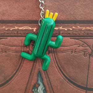 May include: A green cactus keychain with a yellow crown on top. The cactus is in a running pose with arms and legs.