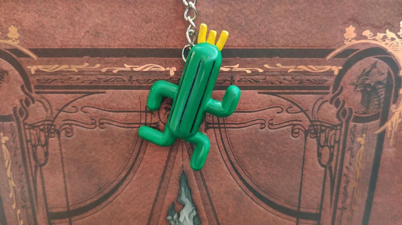 May include: A green cactus keychain with a yellow crown on top. The cactus is in a running pose with arms and legs.