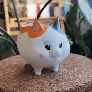 Puede incluir: Una figurita de gato de cer&aacute;mica blanca con marcas naranjas, negras y grises. El gato tiene ojos grandes y un cuerpo redondeado.