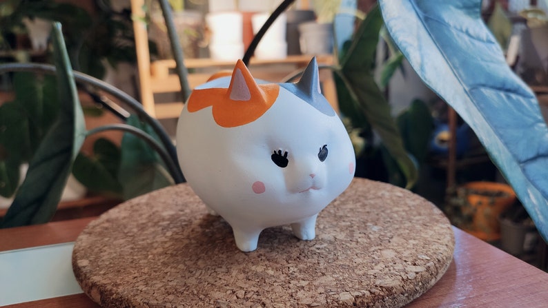Puede incluir: Una figurita de gato de cer&aacute;mica blanca con marcas naranjas, negras y grises. El gato tiene ojos grandes y un cuerpo redondeado.