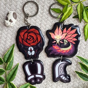 May include: Two keychains with black, red, and white designs. One features a red rose, the other a stylized creature with pink and orange accents. A small cat-shaped charm is also visible. The keychains are attached to silver keyrings.