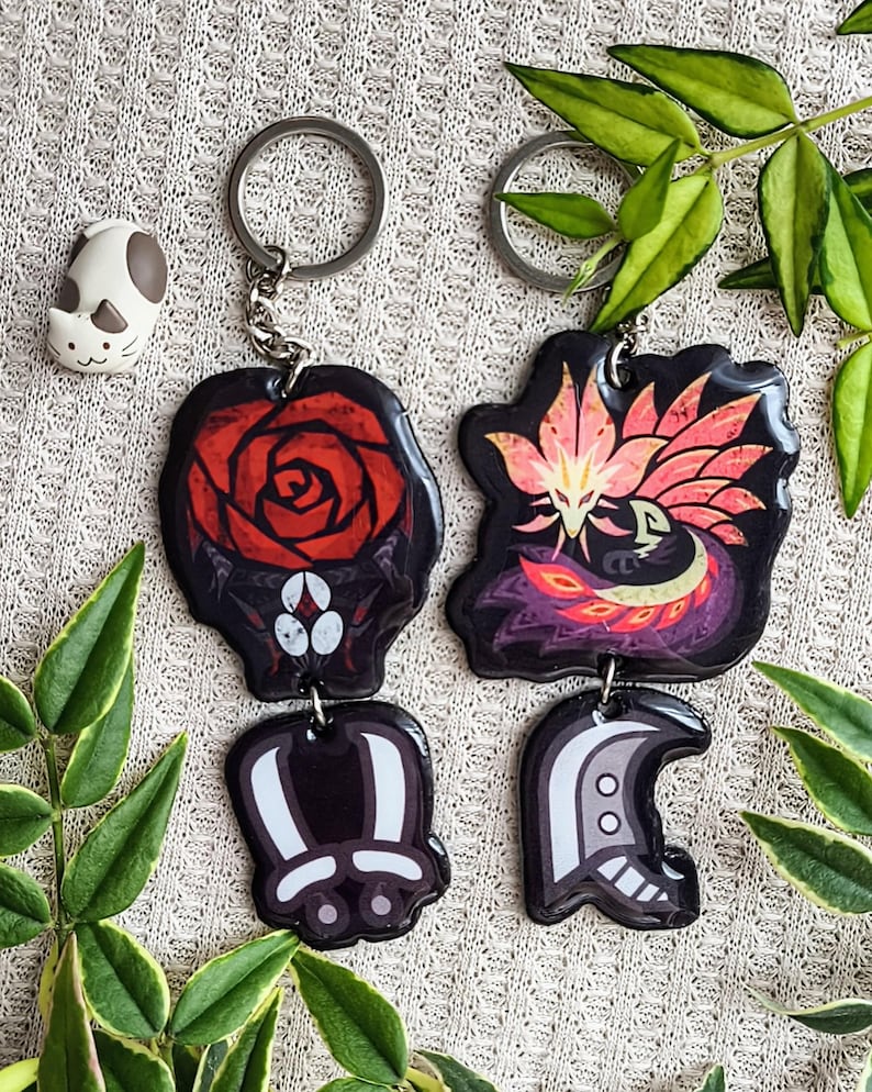 May include: Two keychains with black, red, and white designs. One features a red rose, the other a stylized creature with pink and orange accents. A small cat-shaped charm is also visible. The keychains are attached to silver keyrings.