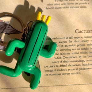 May include: A green Cactuar keychain with yellow spikes. The keychain is made of plastic and has a detailed design. The keychain is hanging from a silver ring.