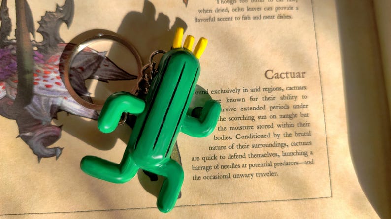 May include: A green Cactuar keychain with yellow spikes. The keychain is made of plastic and has a detailed design. The keychain is hanging from a silver ring.