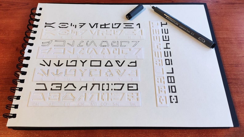 Peut inclure: Un carnet blanc avec une reliure spirale et un stylo noir pos&eacute; dessus. Le carnet comporte plusieurs rang&eacute;es de symboles en relief, peut-&ecirc;tre un alphabet fictif.