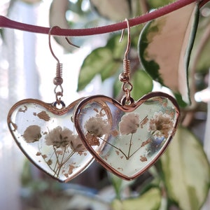 May include: A pair of gold heart-shaped earrings with dried white flowers and gold flakes encased in clear resin.