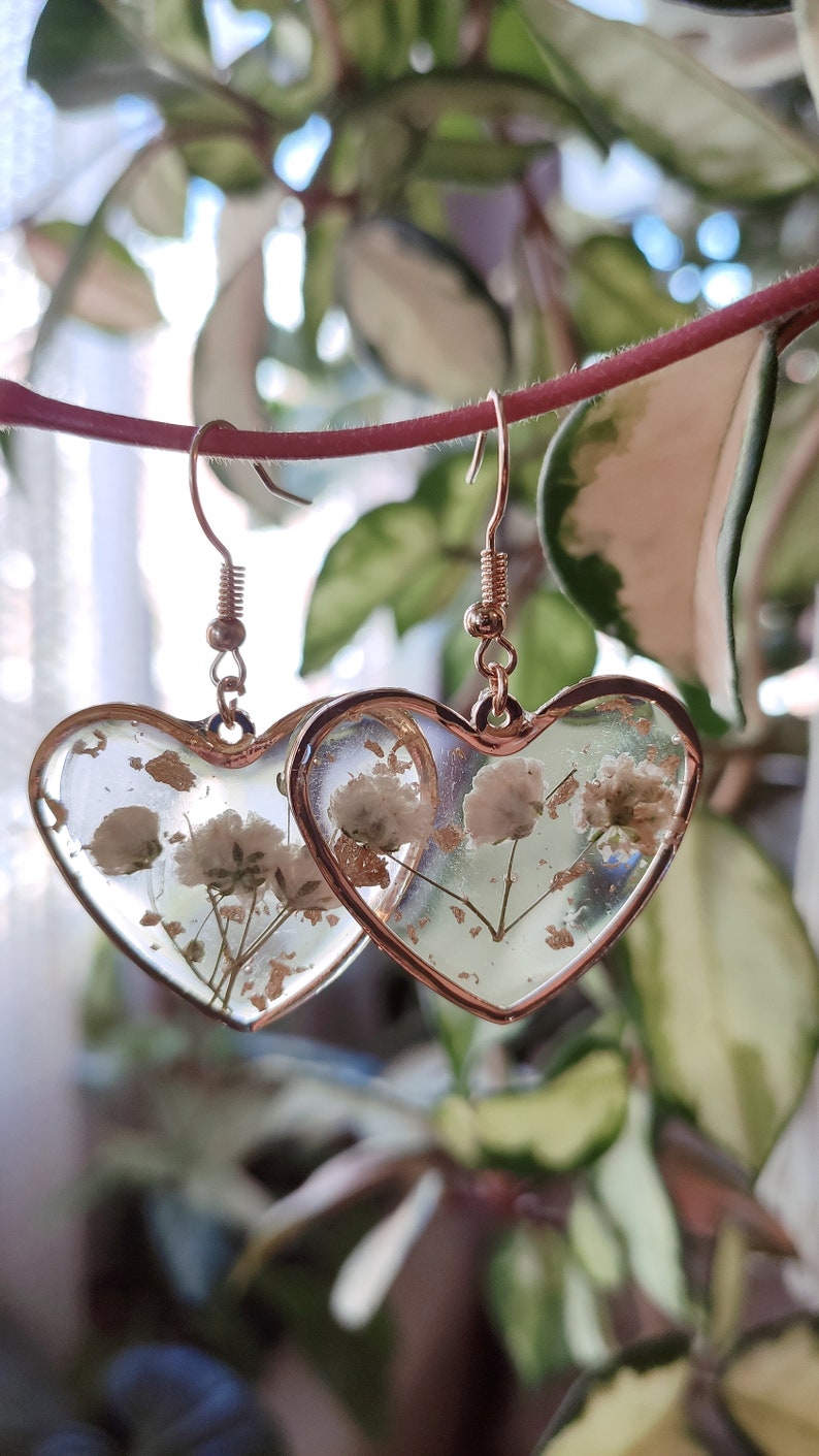 May include: A pair of gold heart-shaped earrings with dried white flowers and gold flakes encased in clear resin.