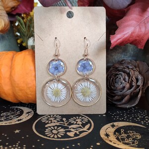 May include: Gold-coloured drop earrings with dried flowers set in clear resin. The earrings have a top section with blue flowers and a bottom section with white daisies. The earrings are displayed on a brown card.