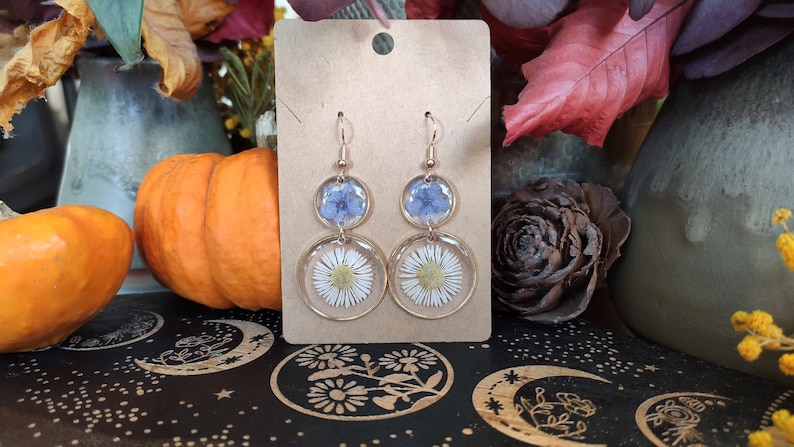 May include: Gold-coloured drop earrings with dried flowers set in clear resin. The earrings have a top section with blue flowers and a bottom section with white daisies. The earrings are displayed on a brown card.
