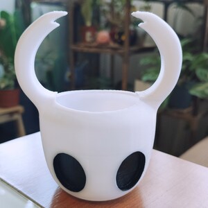 May include: A white ceramic planter shaped like the Knight from the video game Hollow Knight. The planter has two black eyes and two curved horns.