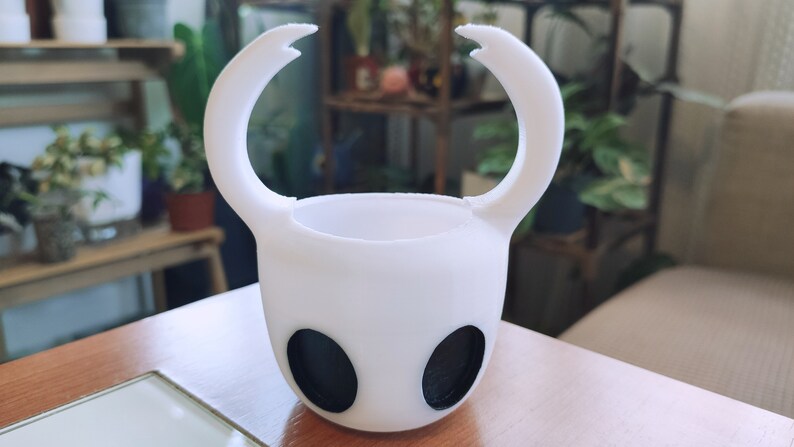 May include: A white ceramic planter shaped like the Knight from the video game Hollow Knight. The planter has two black eyes and two curved horns.