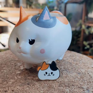 May include: A white ceramic cat planter with orange, black, and gray markings. The planter is sitting on a cork coaster. A small enamel pin of a cat with orange, black, and white markings is in front of the planter.