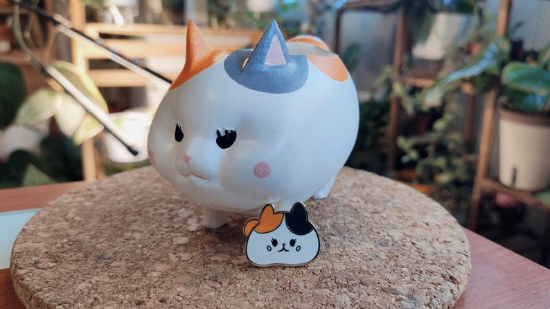 May include: A white ceramic cat planter with orange, black, and gray markings. The planter is sitting on a cork coaster. A small enamel pin of a cat with orange, black, and white markings is in front of the planter.