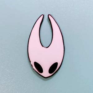 Może przedstawiać: Biała szpilka emaliowana z czarnym obramowaniem, w kształcie pustej maski z dwoma czarnymi oczami. Szpilka jest popularną postacią z gry wideo Hollow Knight.
