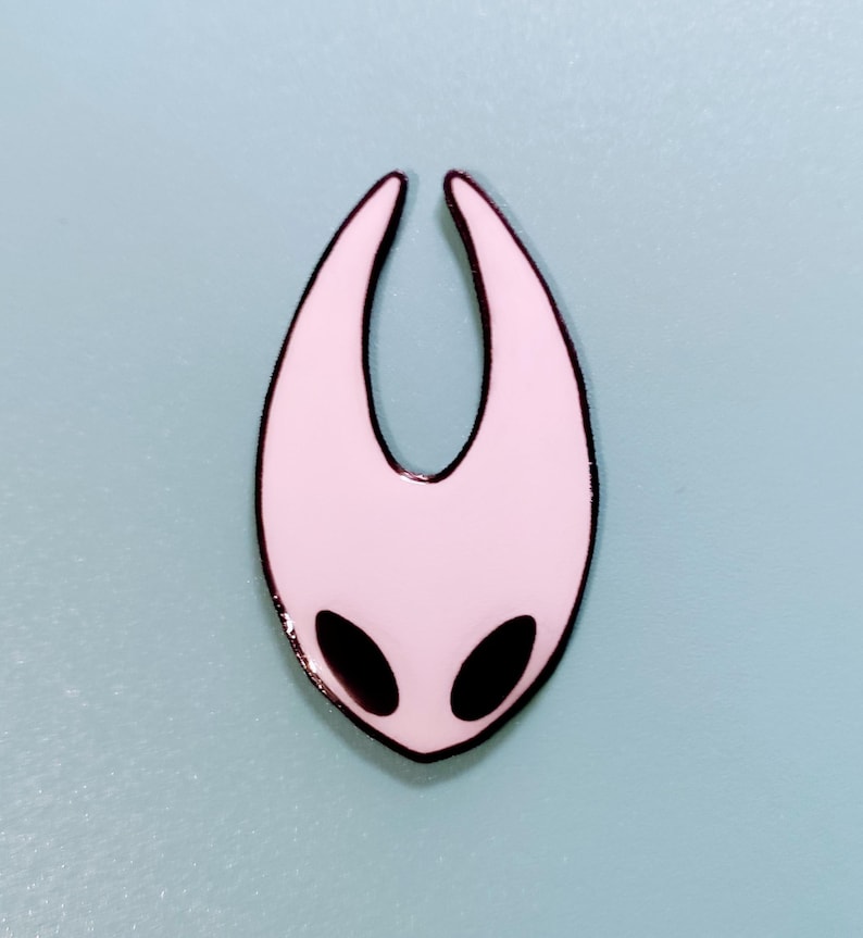 Może przedstawiać: Biała szpilka emaliowana z czarnym obramowaniem, w kształcie pustej maski z dwoma czarnymi oczami. Szpilka jest popularną postacią z gry wideo Hollow Knight.
