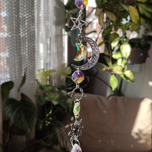 Puede incluir: Un atrapasoles de temática celestial con una cadena plateada. Cuenta con dijes de estrella, luna creciente, planeta y sol, junto con colgantes de cristal iridiscente. El atrapasoles está diseñado para colgar y capturar la luz solar.