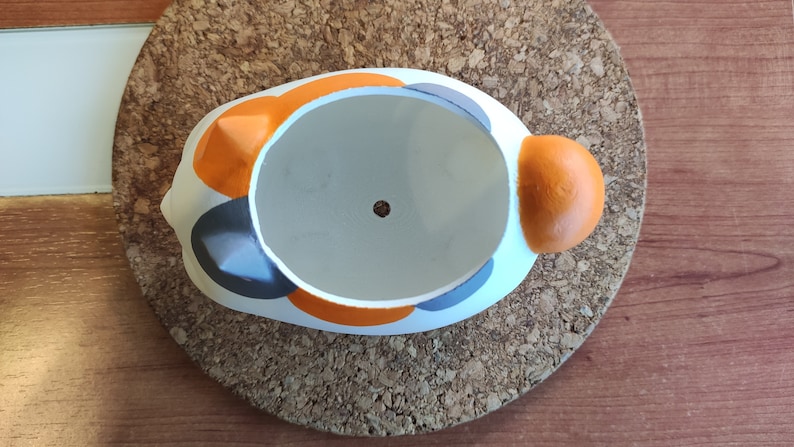Puede incluir: Una maceta de cer&aacute;mica blanca con forma de gato con detalles en naranja, gris y blanco. La maceta tiene un agujero de drenaje en la parte inferior.