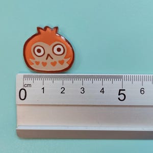 May include: A small orange and brown enamel pin featuring an owl with large eyes. The pin is approximately 2 cm wide and is shown next to a ruler.