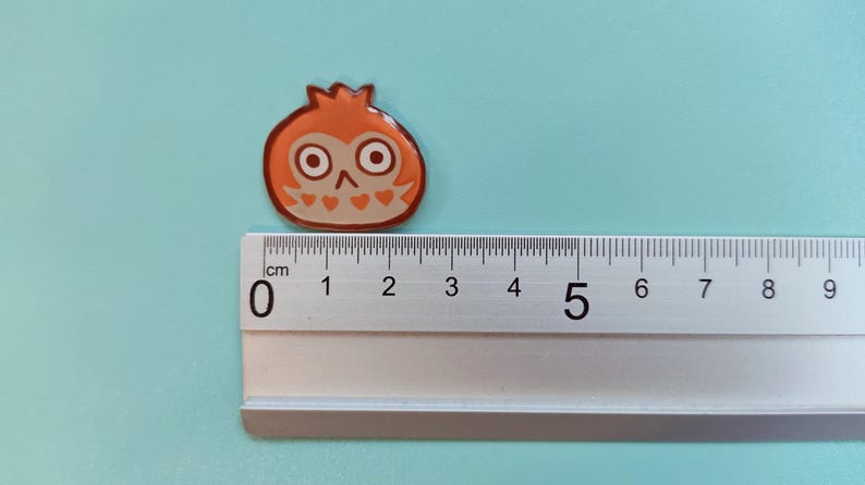 May include: A small orange and brown enamel pin featuring an owl with large eyes. The pin is approximately 2 cm wide and is shown next to a ruler.