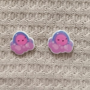 May include: Two cloud-shaped earrings with a sleeping face design. The earrings feature a pink and purple color scheme with a blue outline. The earrings are set against a textured, off-white background.