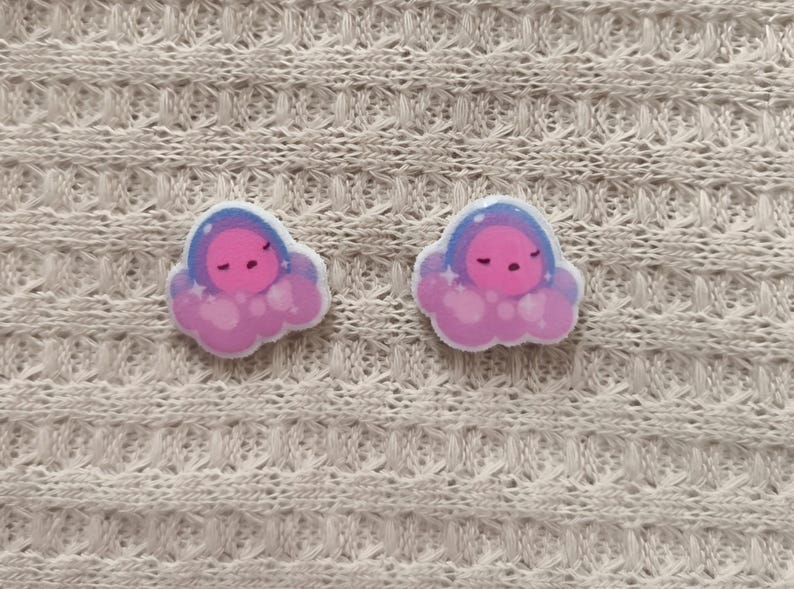 May include: Two cloud-shaped earrings with a sleeping face design. The earrings feature a pink and purple color scheme with a blue outline. The earrings are set against a textured, off-white background.