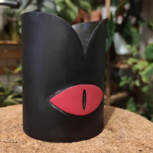 Op de afbeelding: Zwarte plantenpot met een rood oogontwerp. De pot is gemaakt van keramiek en heeft een gladde afwerking.