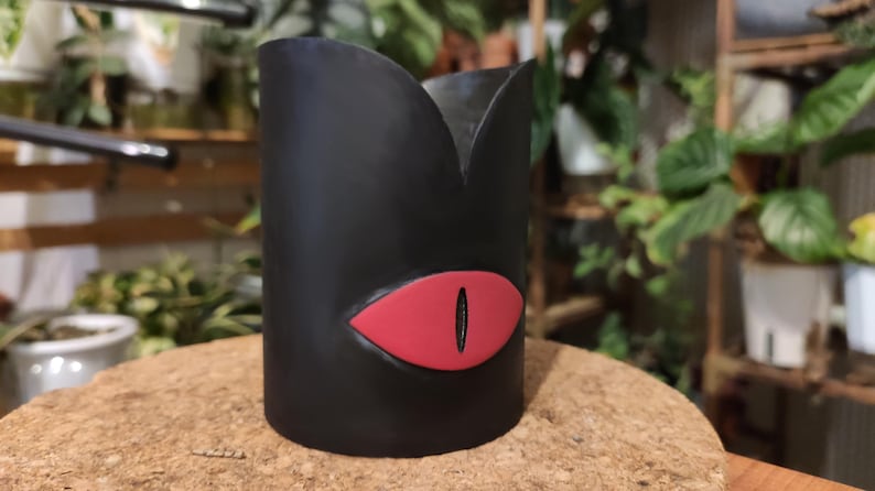 Op de afbeelding: Zwarte plantenpot met een rood oogontwerp. De pot is gemaakt van keramiek en heeft een gladde afwerking.