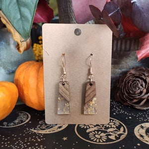 May include: A pair of gold dangle earrings with a rectangular wood and resin design. The resin is clear and contains gold flakes. The earrings are on a brown card.