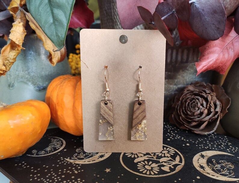 May include: A pair of gold dangle earrings with a rectangular wood and resin design. The resin is clear and contains gold flakes. The earrings are on a brown card.