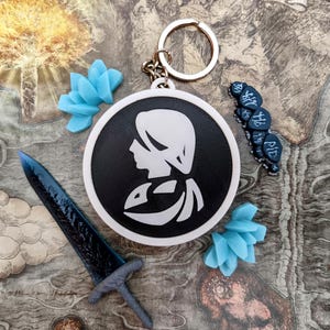 May include: A collection of fantasy-themed accessories, including a keychain with a black and white profile, a blue sword, blue flower-shaped charms, and a string of dark blue beads. The items are arranged on a map-like background.