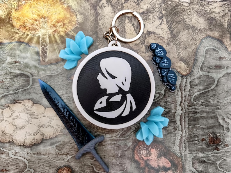 May include: A collection of fantasy-themed accessories, including a keychain with a black and white profile, a blue sword, blue flower-shaped charms, and a string of dark blue beads. The items are arranged on a map-like background.