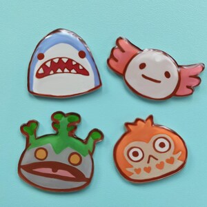 May include: Four colorful enamel pins featuring cartoon animal designs. The pins are shaped like a shark, an axolotl, a creature with green hair, and an owl. The pins are all on a light blue background.