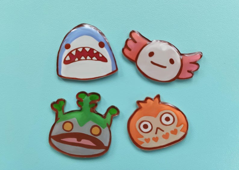 May include: Four colorful enamel pins featuring cartoon animal designs. The pins are shaped like a shark, an axolotl, a creature with green hair, and an owl. The pins are all on a light blue background.