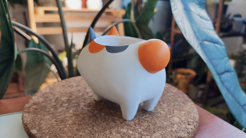 Puede incluir: Maceta de cer&aacute;mica blanca con forma de gato con detalles naranja y negro. La maceta tiene una peque&ntilde;a abertura en la parte superior para plantar.