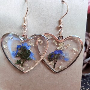 May include: A pair of heart-shaped earrings with gold leaf and blue flowers encased in clear resin. The earrings have gold-toned hooks.