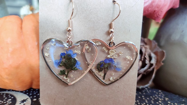 May include: A pair of heart-shaped earrings with gold leaf and blue flowers encased in clear resin. The earrings have gold-toned hooks.