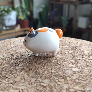 K&ouml;nnte beinhalten: Eine kleine Keramikfigur eines Katers mit wei&szlig;em Fell, orangefarbenen und schwarzen Markierungen und einem runden K&ouml;rper.